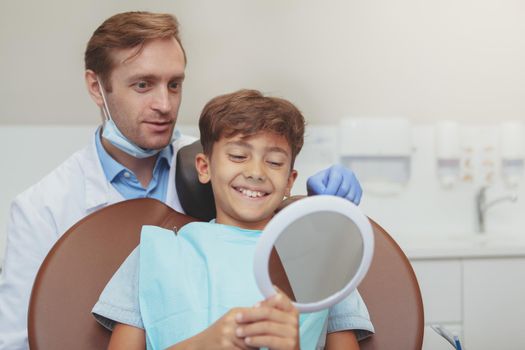 Charming happy little boy smiling, looking in the mirror at the dentists office. Mature male dentist and his young patient checking results of dental care, copy space