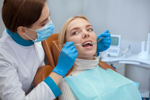 Professional dentist wearing medical mask, examining teeth of a female patient, copy space. Orthodontist working with her client. Beautiful woman having dental checkup. Professionalism concept