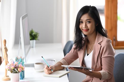 Cheerful asian woman entrepreneur sitting at modern workplace and smiling to camera.