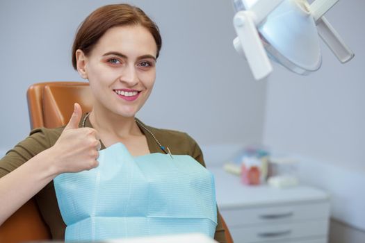 Happy beautiful woman sitting in dental chair, smiling showing thumbs up, copy space. Attractive female patient smiling after successful oral treatment. Happiness, healthcare, dentistry concept
