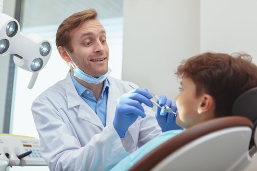 Professional dentist talking to his little patient joyfully, while examining his teeth, copy space. Trustworthy doctor concept