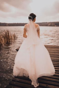 A young woman bride in white wedding dress red curly hair stands on wooden brown pier in middle of blue azure lake.