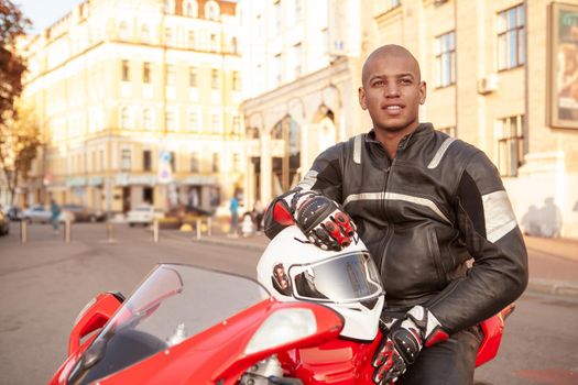 Attractive young African man looking around in the city, sitting on his motorbike. Handsome male biker travelling Europe on his motorcycle