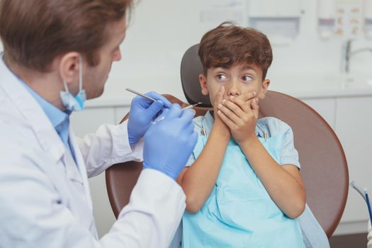 Little terrified boy covering his mouth with his hands, sitting in a dental chair. Cropped shot of a professional dentist trying to examine teeth of a scared little boy