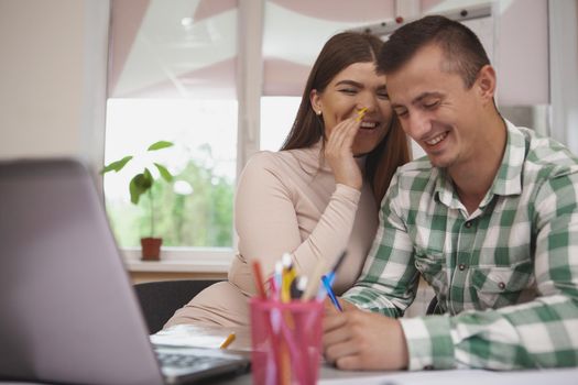 Love, education concept. Charming young couple studying together at college library, using laptop. Beautiful cheerful female student whispering something funny to her boyfriend