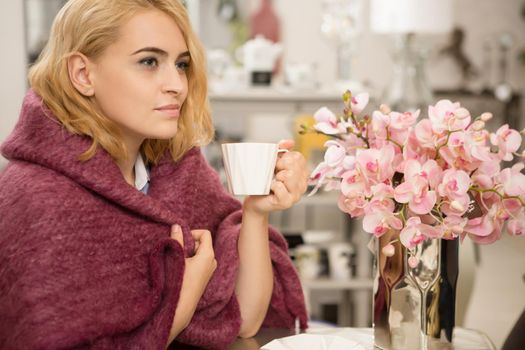 Attractive blonde young woman enjoying cup of tea or coffee sitting wrapped in a woolen warm blanket relaxing at home on a weekend copyspace lifestyle relaxation harmony femininity beauty leisure