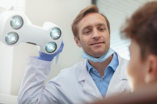 Cropped shot of a professional dentist adjusting dental lamp before examining teeth of a young boy. Concentrated dentist preparing for dental checkup with his patient