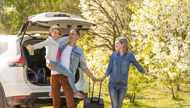 Happy family standing together near a car with open trunk enjoying view of rural landscape nature. Parents and their kid leaning on vehicle luggage compartment. Weekend travel and holidays concept