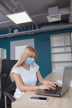 Vertical shot of a female entrepreneur working on laptop, wearing protective face mask at the office