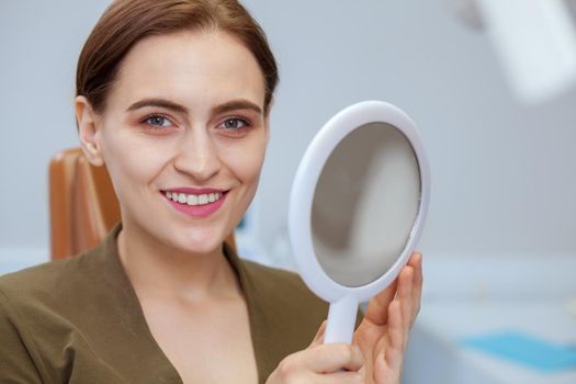 Close up of a beautiful woman smiling happily to the camera, after checking out her perfect healthy teeth in a mirror. Cheerful female patient at the dental clinic, copy space