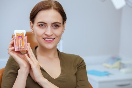 Young cheerful female smiling to the camera posing at dental clinic with a tooth model in her hands. Attractive woman holding tooth mold, sitting in dental chair, copy space. Medicine, service