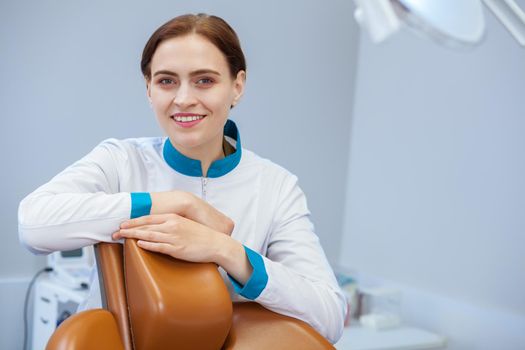 Lovely young woman doctor posing at dental office, smiling joyfully to the camera. Female dentist at her clinic. Cheerful orthodontist ready for patients, copy space. Happiness, health concept