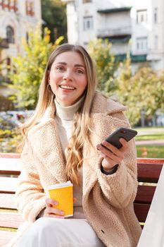 Vertical shot of a beautiful happy woman having coffee in the park in autumn, using her smart phone