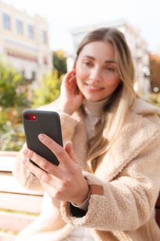 Selective focus on smart phone in the hand of a beautiful happy woman outdoors on sunny autumn day