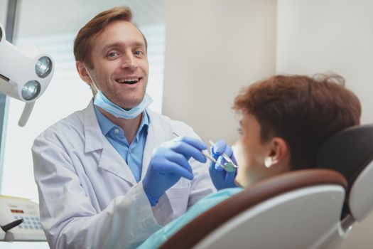 Cheerful mature male dentist enjoying working at his clinic, examining teeth of a little boy, copy space