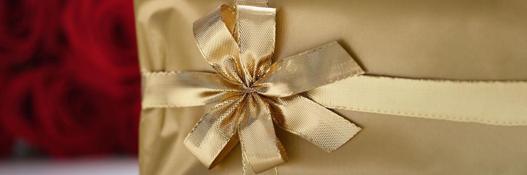 Gift box in gold packaging with ribbon, close-up on a background of red roses. Romance and pleasant surprise