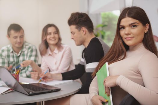 Beautiful young female student smiling to the camera, her classmates working on the background, copy space. Cheerful lovely woman enjoying working on a project with her college friends