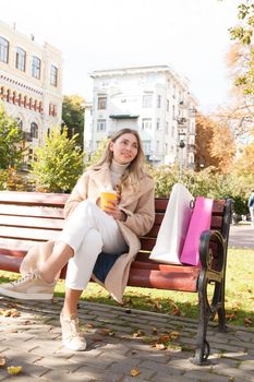 Vertical full length shot of a beautiful woman sitting in the park with her shopping bags and cup of coffee