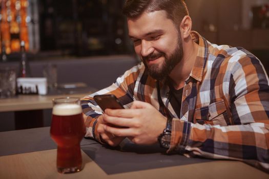 Happy handsome man texting on his smart phone at beer pub. Attractive cheerful man using his phone while drinking beer at the bar