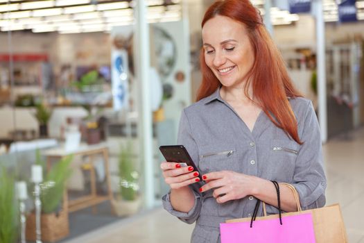 Cheerful woman using her smart phone at the shopping mall, copy space. Attractive female customer walking with shopping bags at the mall, browsing online on her phone