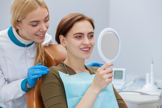 Young woman checking out her healthy white teeth in the mirror, dentist standing near, copy space. Professional dentist and her client examining results of medical teeth treatment. Dentistry concept