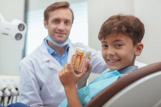 Dental health, children concept. Close up of a cheerful handsome young boy smiling joyfully, holding tooth model sitting in a dental chair. Little boy visiting dentist for a checkup