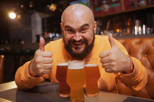 Excited bearded man laughing, showing thumbs up sitting in front of three beer glasses at the pub