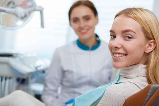 Gorgeous happy woman with perfect healthy teeth smiling to the camera, sitting at dental chair, copy space. Cheerful woman and her dentist smiling joyfully after examination at the clinic. Dentistry