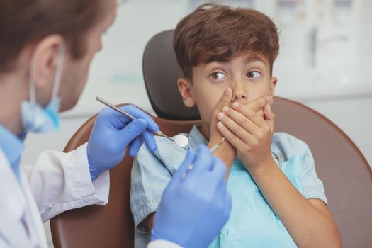 Scared child, dentistry, toothache concept. Cropped close up of a young boy hiding his mouth behind hands, refusing to get teeth checkup by professional dentist.