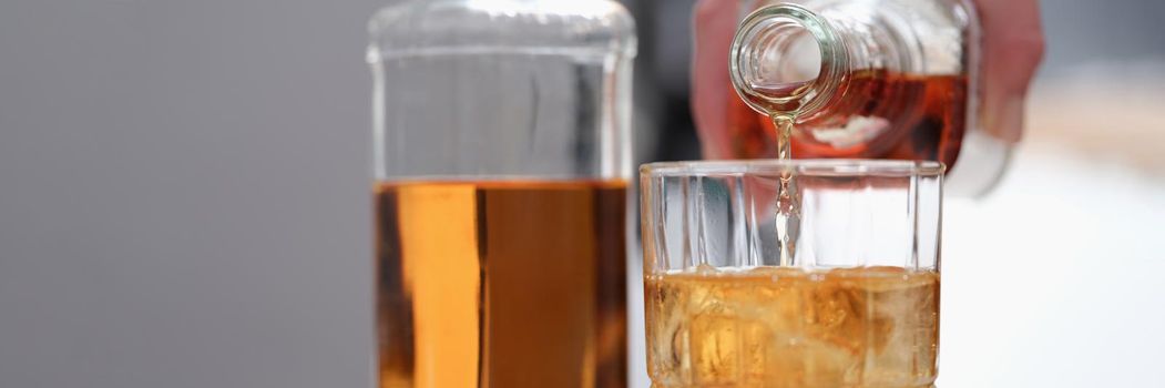 A hand pours alcohol from a bottle into a beautiful glass, close-up. Whiskey with ice, alcoholic cocktail, party