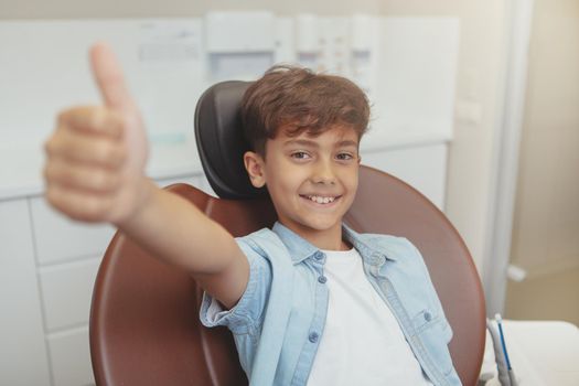 Lovely cheerful young boy smiling, showing thumbs up sitting in a dental chair. Adorable little boy looking happy after dental examination at the clinic