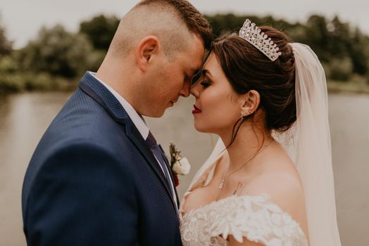 blonde European Caucasian young man groom in blue suit and black-haired woman bride in white wedding dress with long veil and tiara on head. Newlyweds hold hands smile kiss and look at each other near river