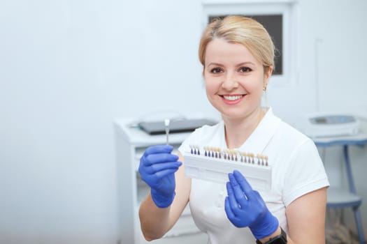 Charming female dentist smiling to the camera, holding teeth whitening shade chart, copy space
