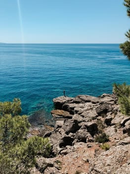 Old man hobby fishing angling standing on big rock stone. senior pensioner catches fish spinning. Top view Montenegro Sutomore stones beach turquoise Adriatic sea water mountains. Summer background