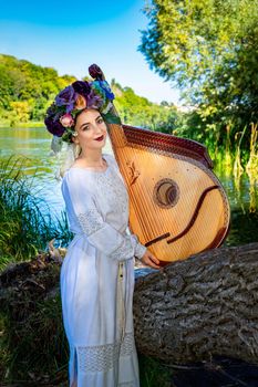 Young Ukrainian woman in national dress plays a bandura by the river on a summer sunny day.