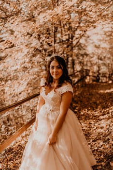 woman bride in white wedding dress with hairstyle makeup and crown on her head walks through autumn forest on fallen orange leaves