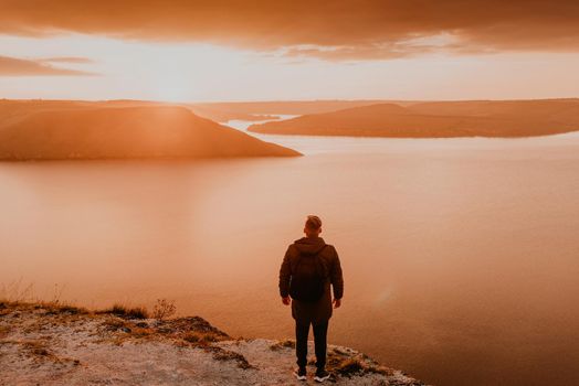 man in casual clothes stands on mountain. fiery orange sunset sunset rays through clouds. panoramic view from cliff to river lake sea in distance silhouettes islands