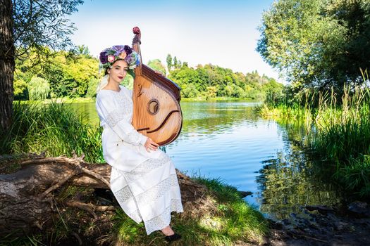 A young Ukrainian woman in national dress and a wreath on her head is sitting by the river with a bandura in her hands.