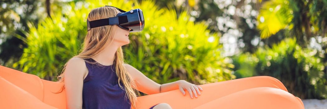 BANNER, LONG FORMAT Summer lifestyle portrait of pretty girl sitting on the orange inflatable sofa and uses virtual reality headset on the beach of tropical island. Relaxing and enjoying life on air bed.