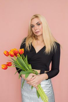 Fashion model woman in fashionable clothes on pink background. Wearing stylish clothing, black blouse, silver skirt. Posing in studio. Holding red tulips in her hands