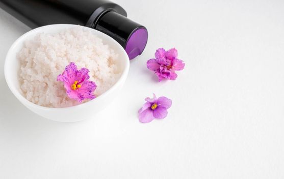 An oval white bowl with pink salt stands on a white background next to it are a black bottle and violet flowers.