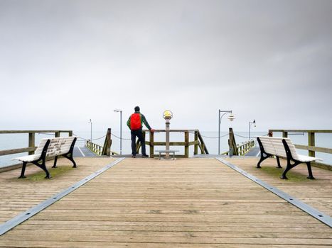 Tourist with red backpack on wooden pier above sea. Man in trekking suit in harbor in dark rainy day. Touristic mole, wet wooden floor above sea. 