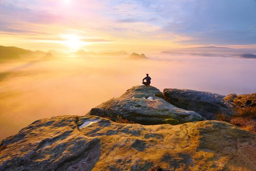 Hiker in squatting position on peak of rock and watching into colorful mist and fog in morning valley. Orange daybreak at horizon