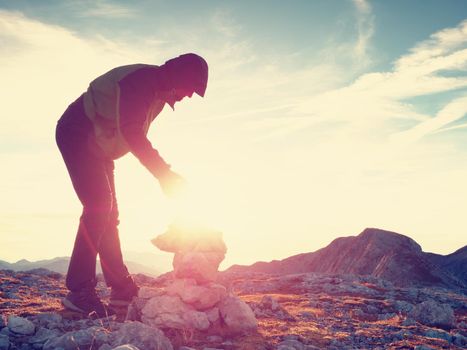 Man put the last stone intopeak of  pyramid. Balanced stone pyramid on  mountain summit. Blue sky with sunset  above mountain. Poor lighting conditions.