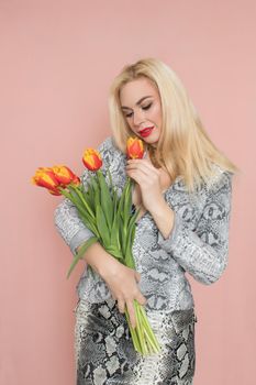 Fashion photo of a beautiful elegant young woman in pretty snake suit, jacket blazer, top, skirt, massive chain around the neck posing on pink background. Studio shot. Bouquet orange tulips holding in hands.