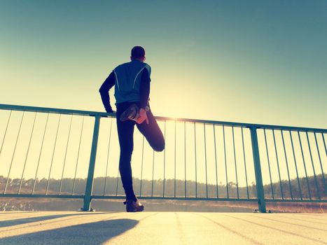 The runner in tall black leggings makes body stretching on bridge path. Sun is outlining man body. Outdoor exercising on smooth concrete ground on lake bridge. 