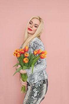 Fashion photo of a beautiful elegant young woman in pretty snake suit, jacket blazer, top, skirt, massive chain around the neck posing on pink background. Studio shot. Bouquet orange tulips holding in hands.