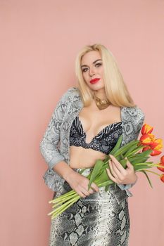 Fashion photo of a beautiful elegant young woman in pretty snake suit, jacket blazer, top, skirt, massive chain around the neck posing on pink background. Studio shot. Bouquet orange tulips holding in hands.