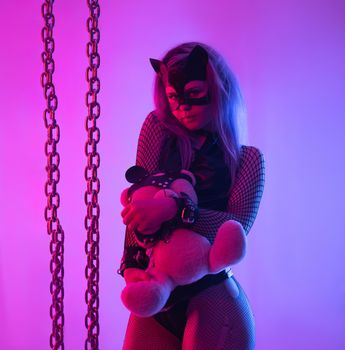 the Sexy girl in latex dress bdsm mistress and cat mask in neon light with chains and teddy bear in leather straps