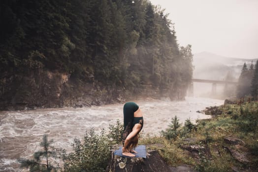 A young blonde woman practices yoga on nature on a stone near a raging river. Green tracksuit.